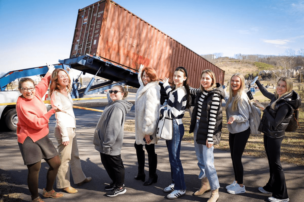 A shipping container being taken off a truck in the background. A group of women are in front of it pointing at the container.